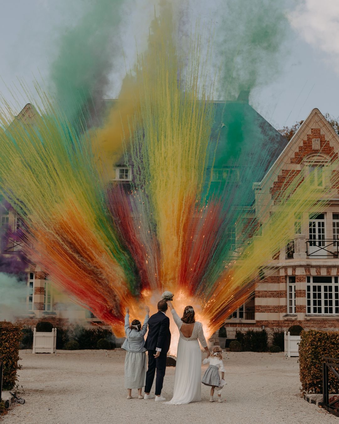 Couple de mariés et leurs enfants contemplant un nuage de fumées orangées et colorées d'un feu de jour sur mesure devant un château, réalisé par Wagnon Group.