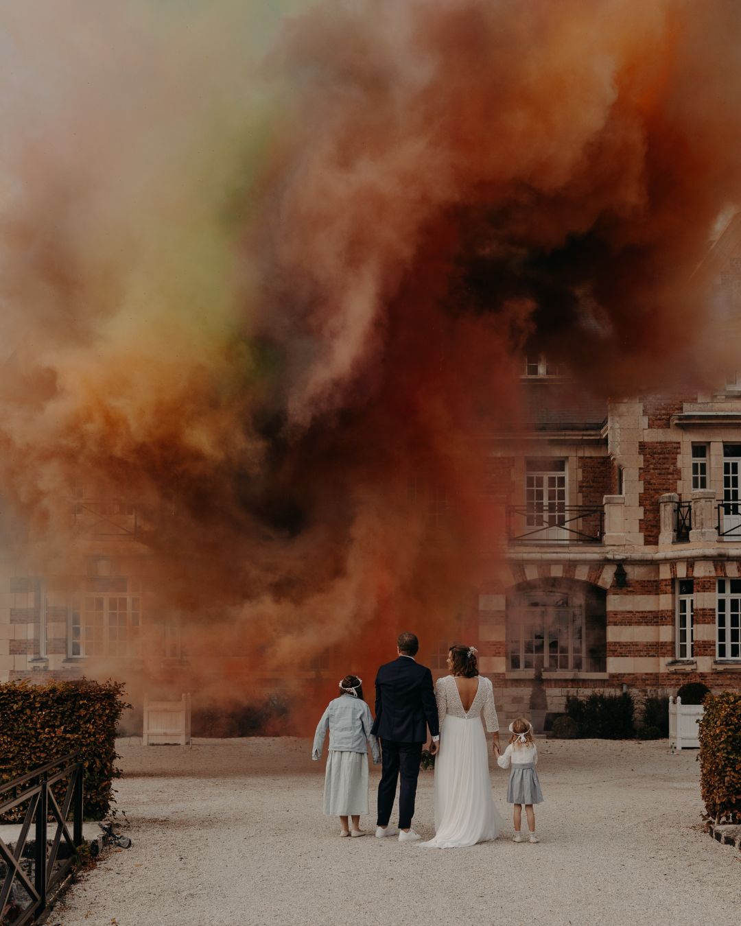 Couple de mariés et leurs enfants bras levés devant une explosion pyrotechnique de jour aux couleurs arc-en-ciel — rouge, vert, jaune, orange — devant un château, spectacle feu de jour sur mesure réalisé par Wagnon Group.