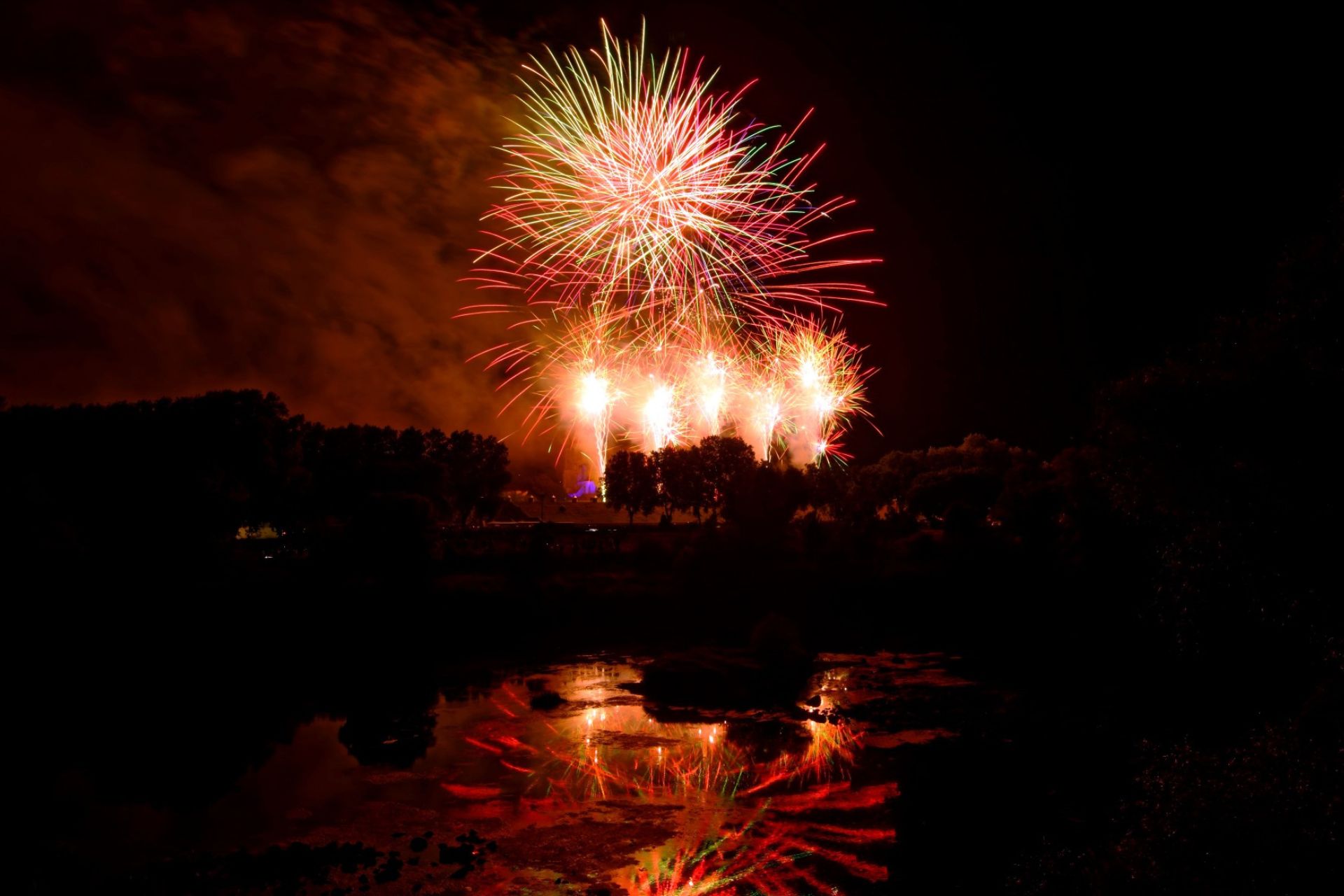 Vue panoramique du spectacle pyrotechnique du 14 juillet à Toul avec reflets des feux d'artifice sur l'eau, silhouette de la cathédrale Saint-Étienne visible en arrière-plan, réalisé par JSE société de Wagnon Group.