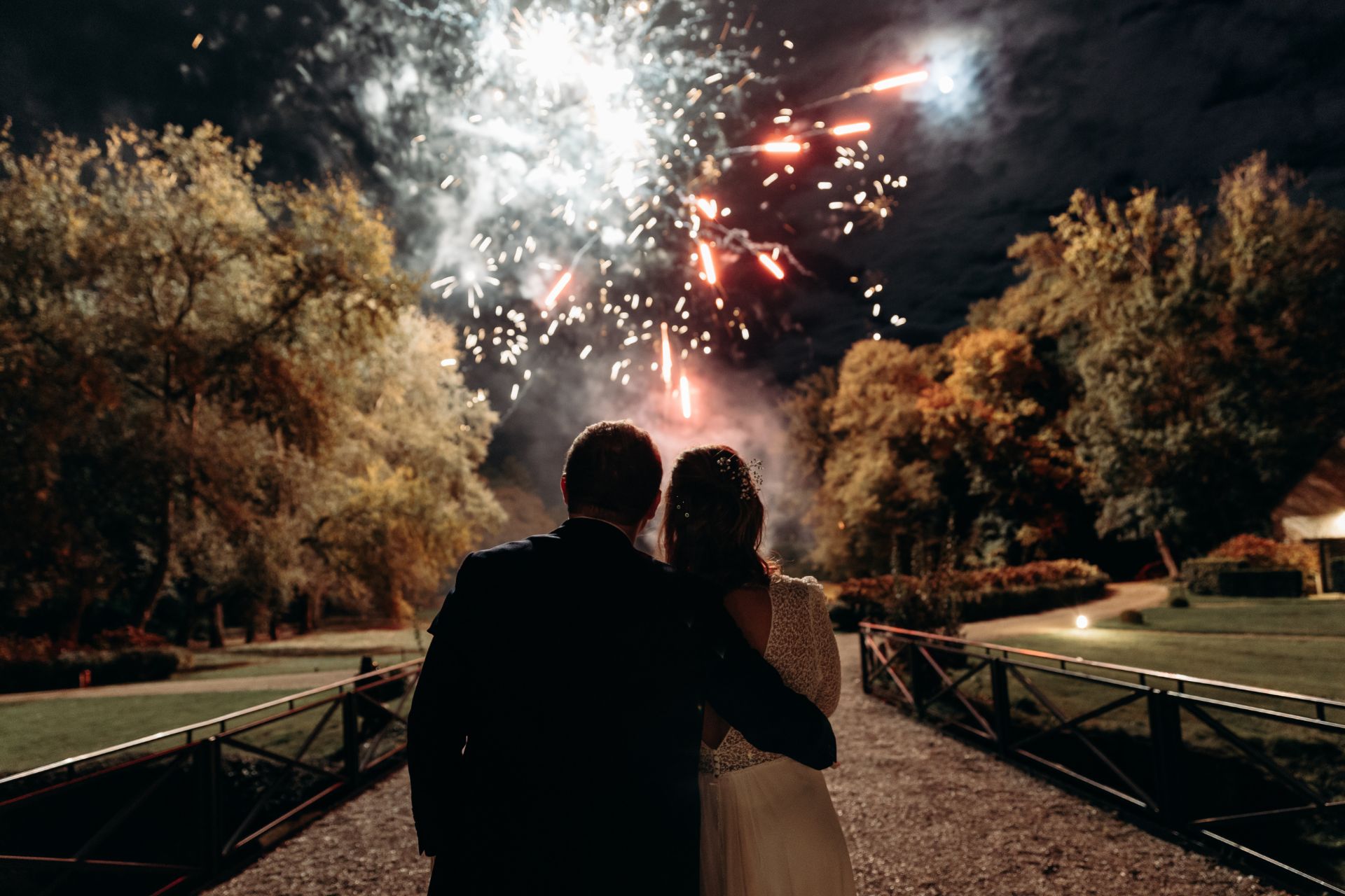 Couple de marié admirant le feu d'artifice blanc et rouge s'élevant dans le ciel nocturne au-dessus d'un parc arboré, spectacle sur mesure réalisé par Wagnon Group.