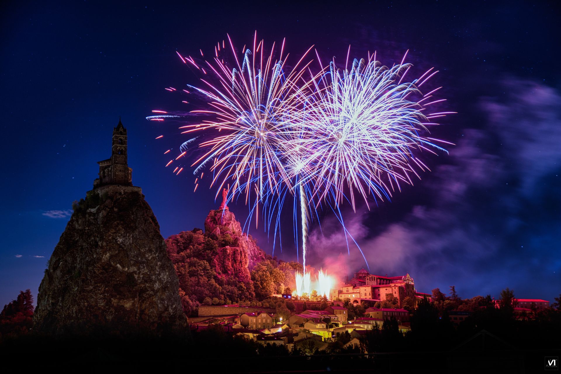 Bouquet de feux d'artifice bleu, blanc et rose explosant dans le ciel nocturne au-dessus du rocher Saint-Michel d'Aiguilhe et de la statue Notre-Dame de France au Puy-en-Velay en Haute-Loire, spectacle pyrotechnique du 14 juillet réalisé par Arsotec société de Wagnon Group en Auvergne-Rhône-Alpes.