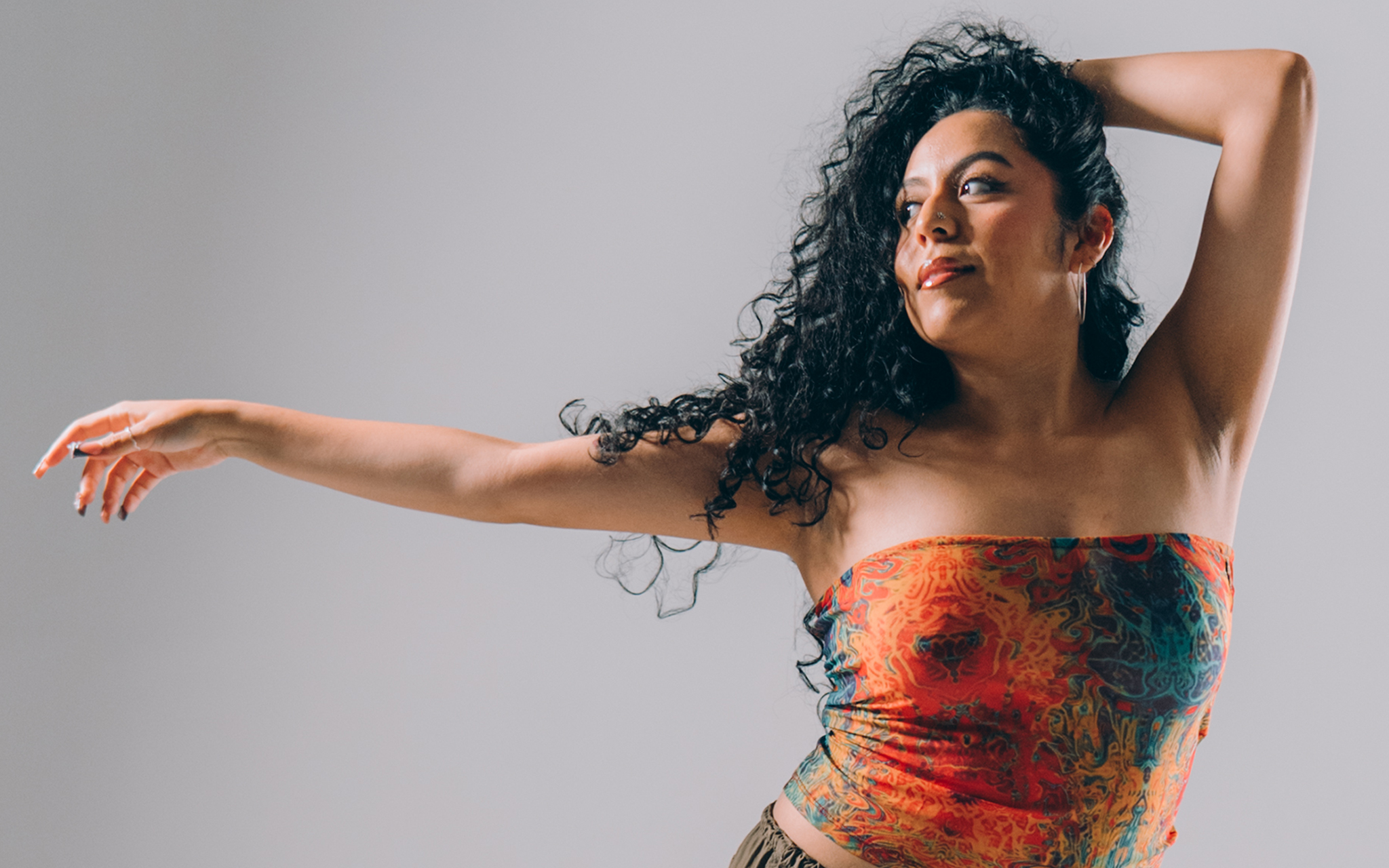 A person poses with one arm extended and one hand behind their head against a plain background.