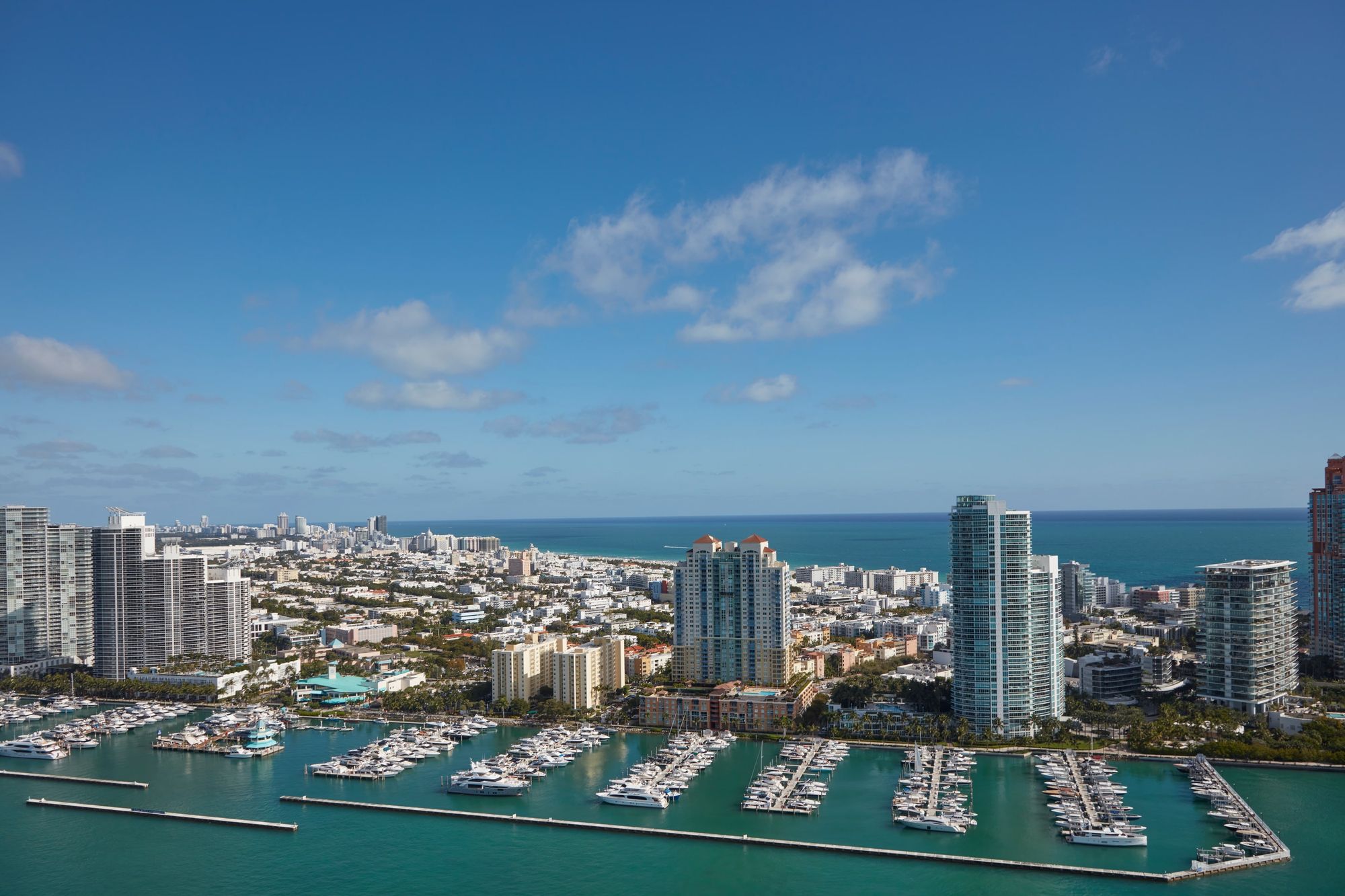Marina with numerous boats docked, surrounded by high-rise buildings along a coastline under a blue sky with scattered clouds.