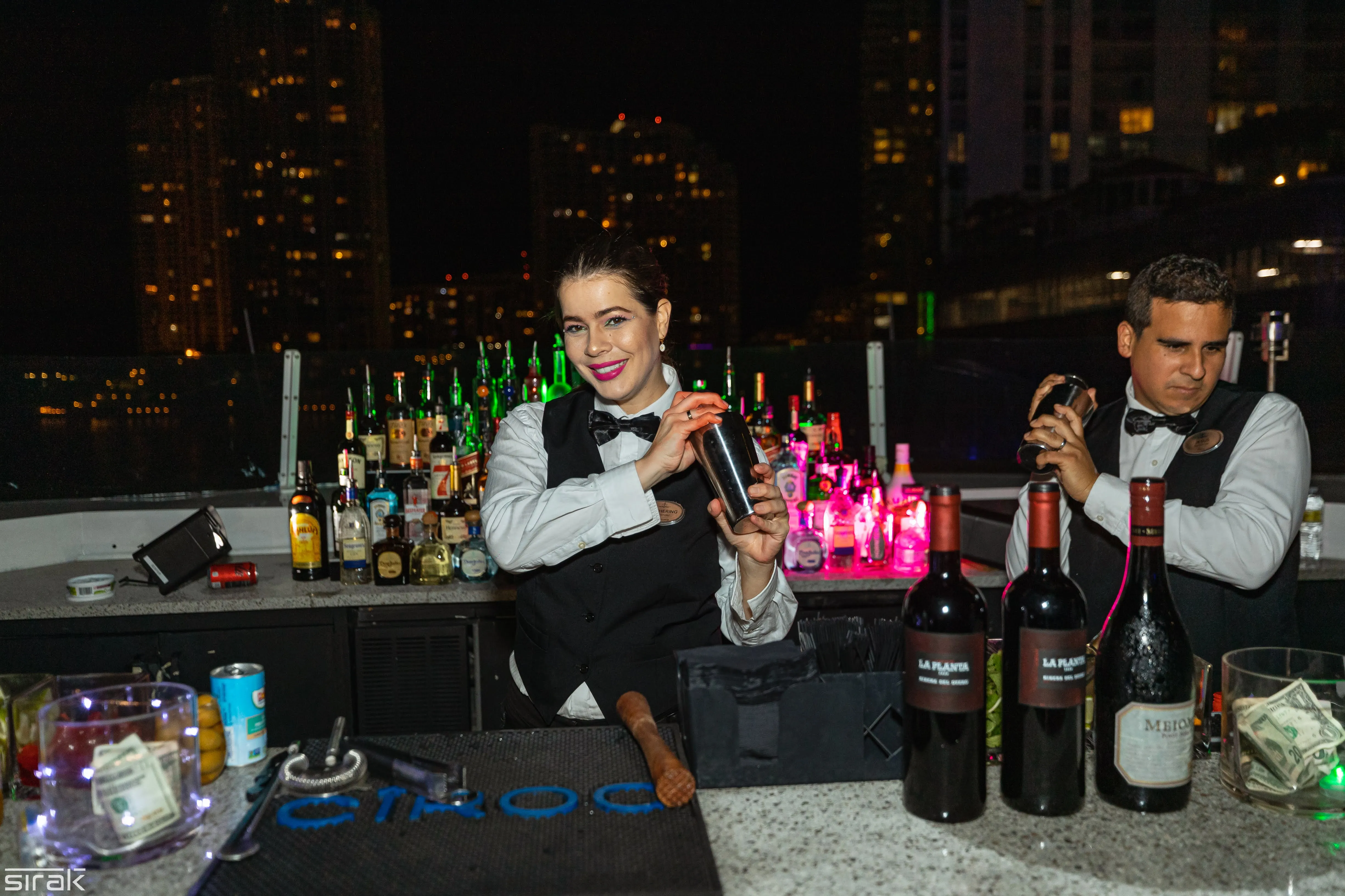 Bartenders at the open-air deck bar at night, Miami skyline behind