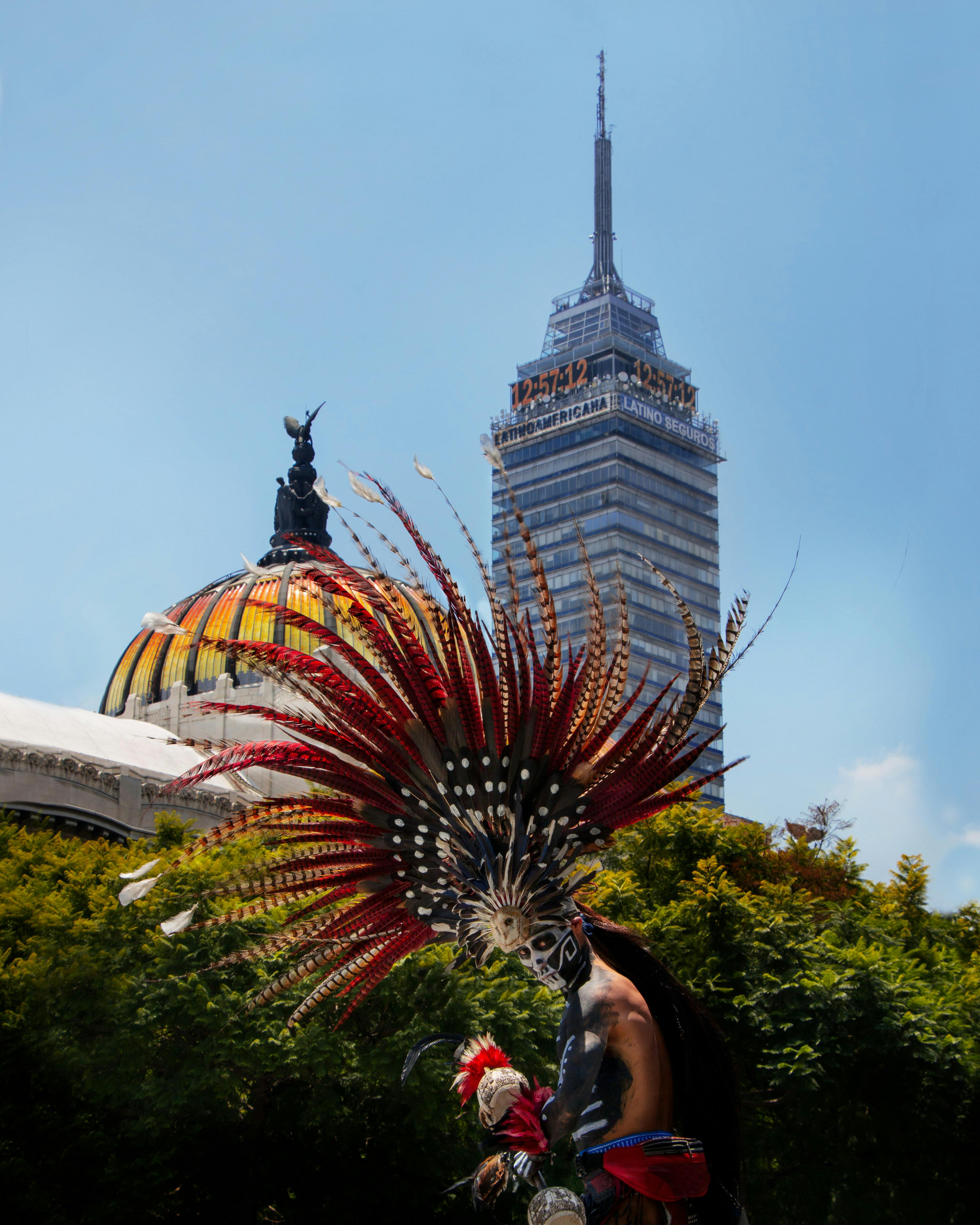 Person in traditional Aztec attire with feathered headdress and face paint performing in front of the Palacio de Bellas Artes dome and Torre Latinoamericana in Mexico City.