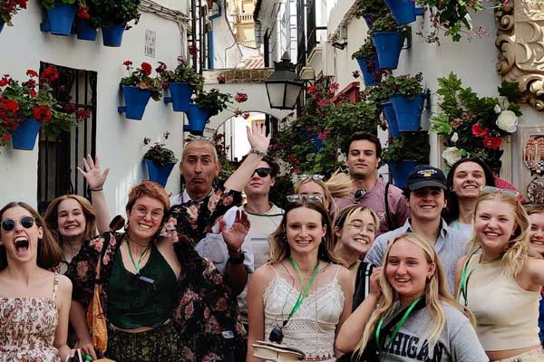 Group of smiling young adults posing together outdoors on a street decorated with blue flower pots and red flowers on white walls.