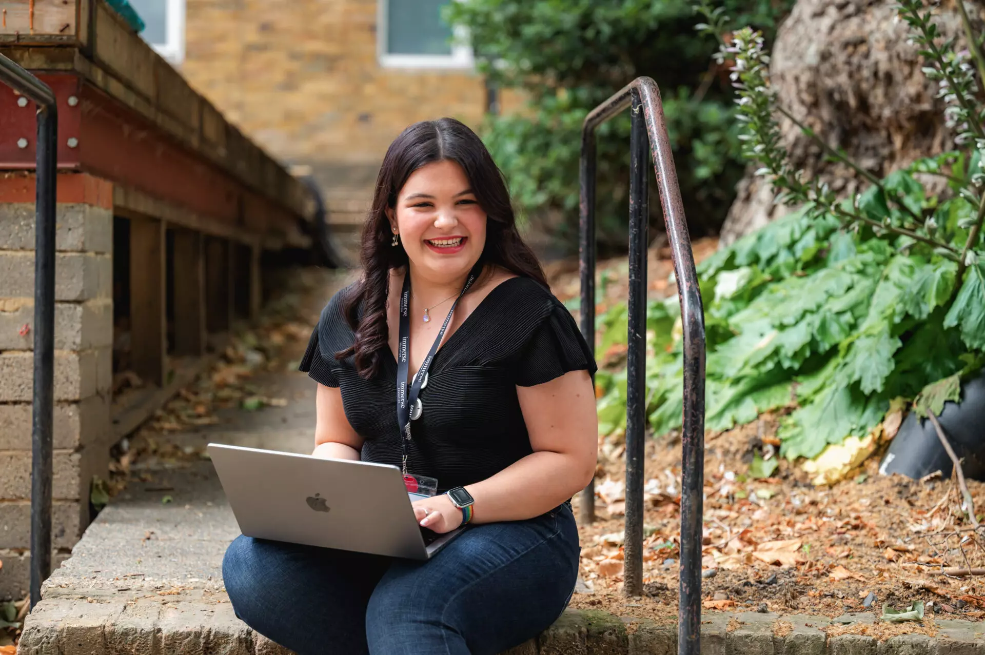 Smiling woman sitting outdoors on a step using a MacBook laptop.