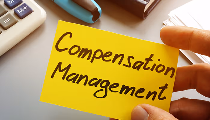 Hand holding a yellow note that reads “Compensation Management” beside office supplies
