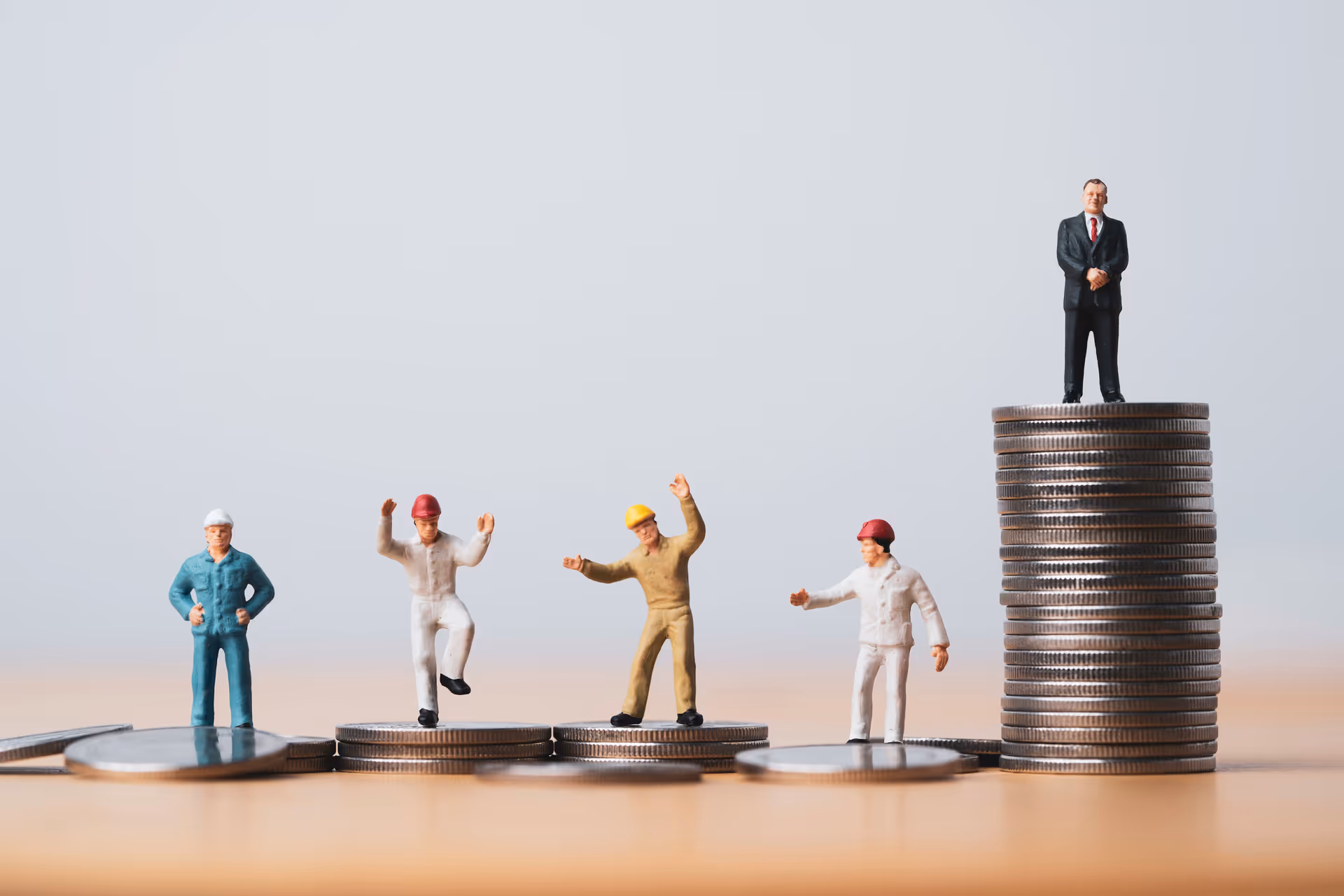 Mini worker figures stand on stacks of coins at different heights, showing income gaps, while a suited figure stands on the highest stack