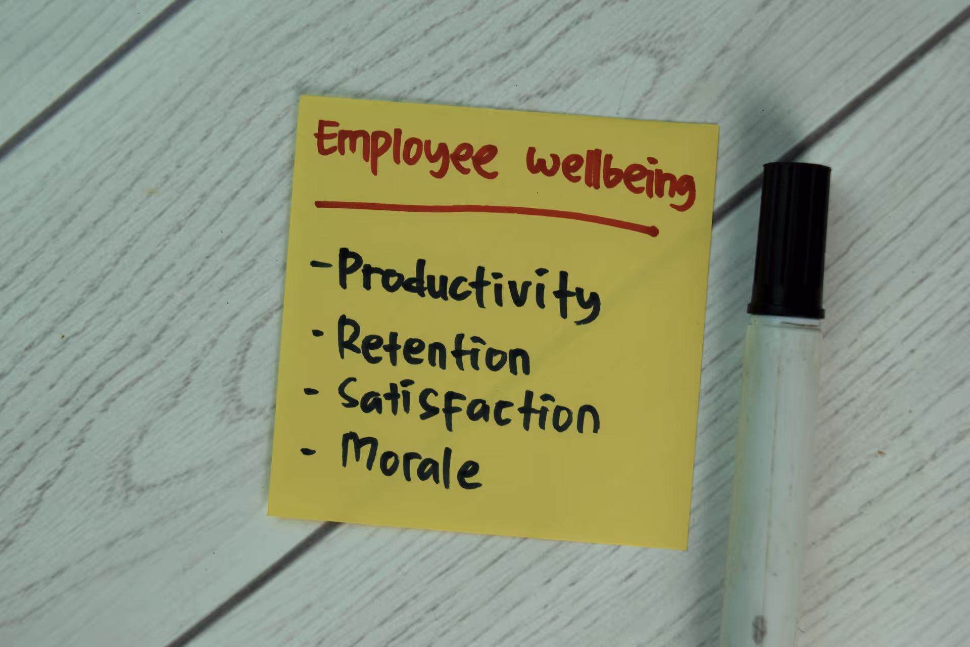 Sticky note titled employee wellbeing listing productivity, retention, satisfaction, and morale with a marker beside it