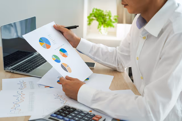 Person reviewing printed charts at a desk with a laptop and calculator, analyzing data and financial insights