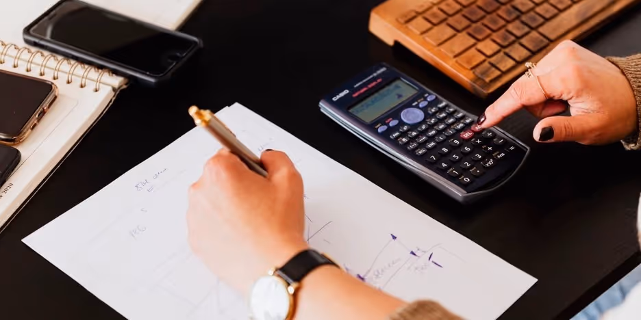 Person using a calculator and writing on paper at a desk with notebooks and a keyboard