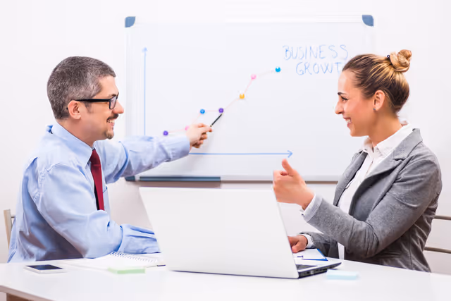 Two colleagues discussing a rising business growth chart on a whiteboard, collaborating with a laptop in an office