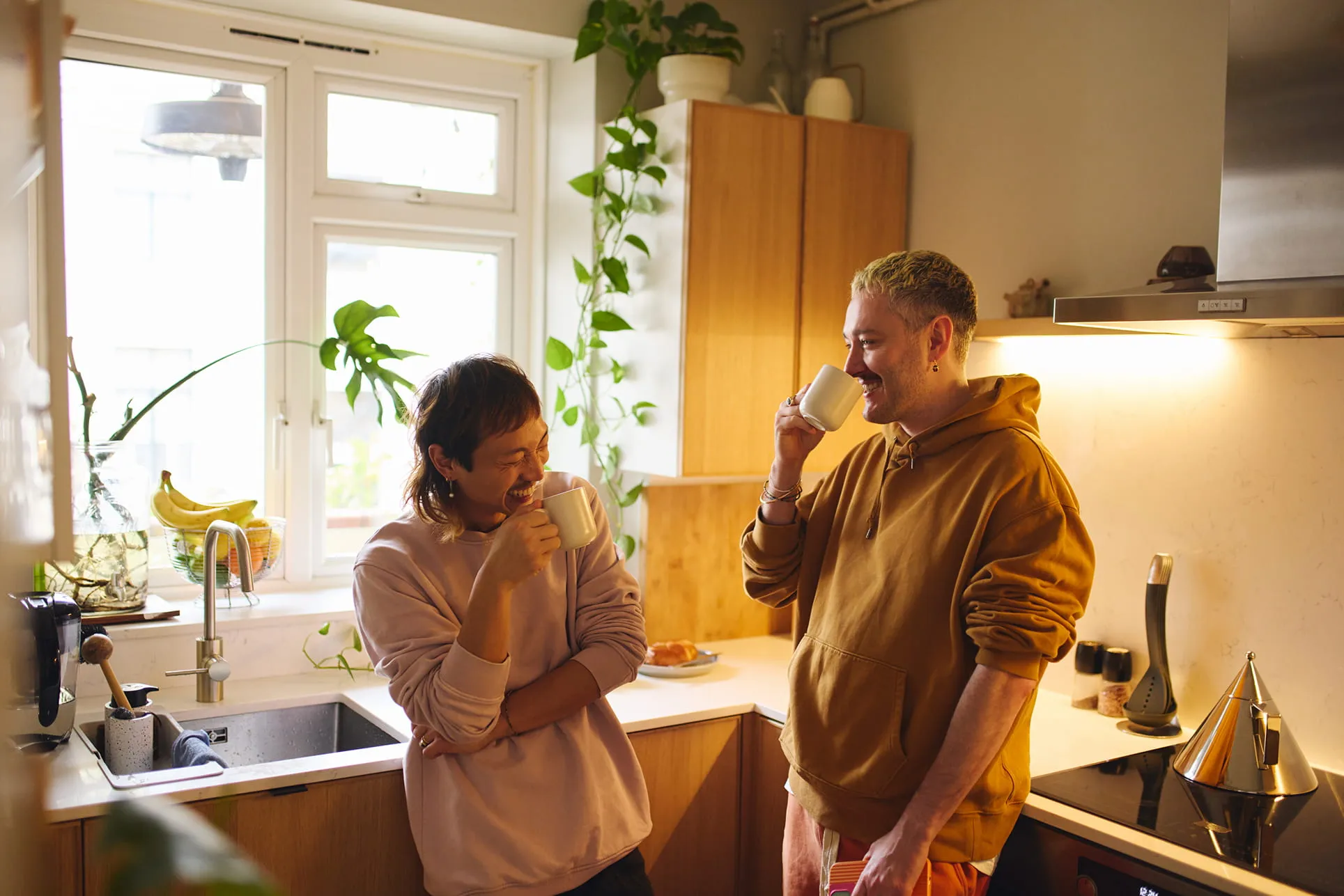 Two people smiling and drinking from white mugs while standing in a cozy kitchen with wooden cabinets and indoor plants.