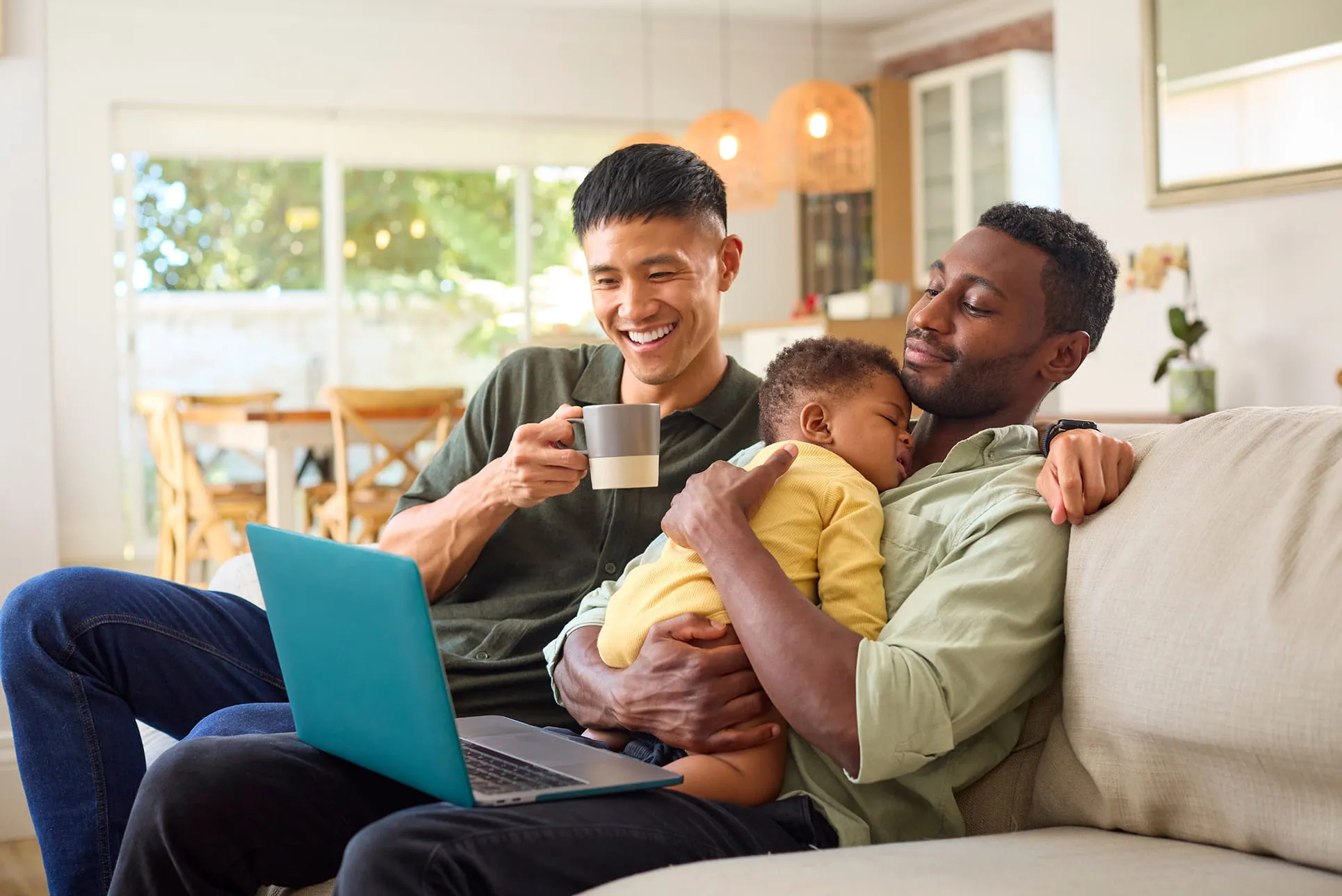 Two men sitting on a couch with a laptop, one holding a sleeping child and the other holding a coffee cup.