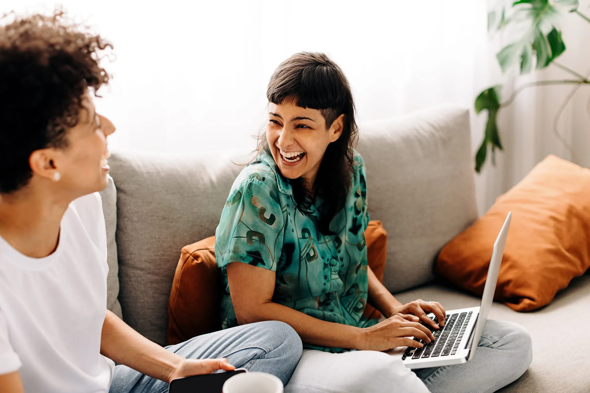 Two women sitting on a couch, one typing on a laptop and both smiling and laughing.