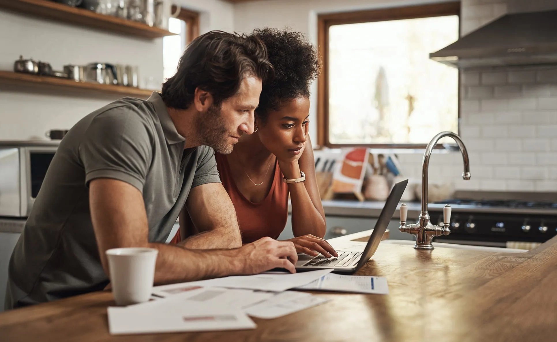 Couple focused on laptop screen in kitchen, reviewing papers on wooden countertop.