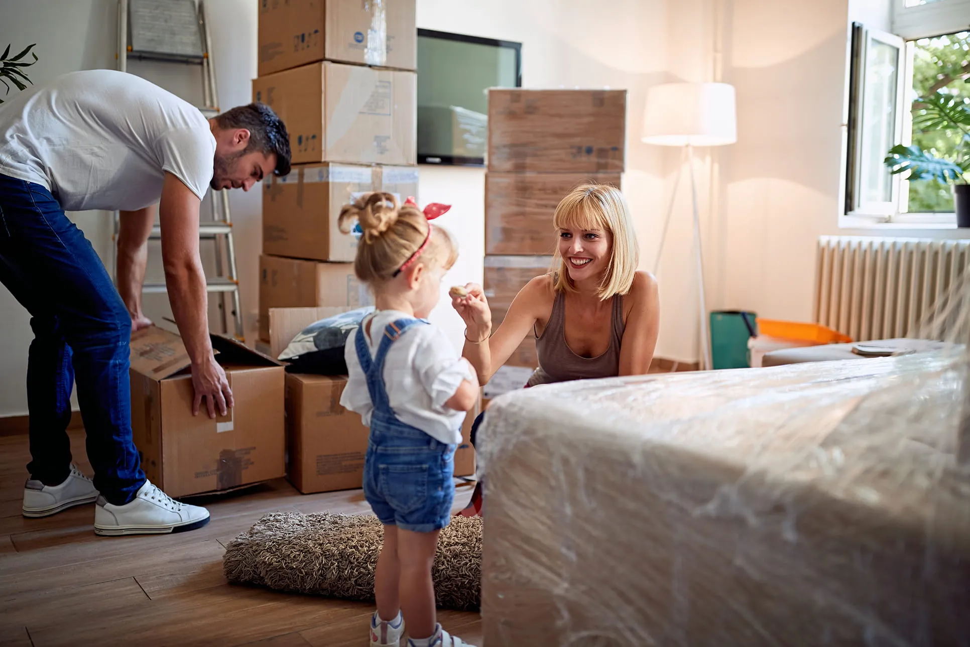 Smiling woman handing a cookie to a young girl in a room full of packed moving boxes while a man opens a box in the background.