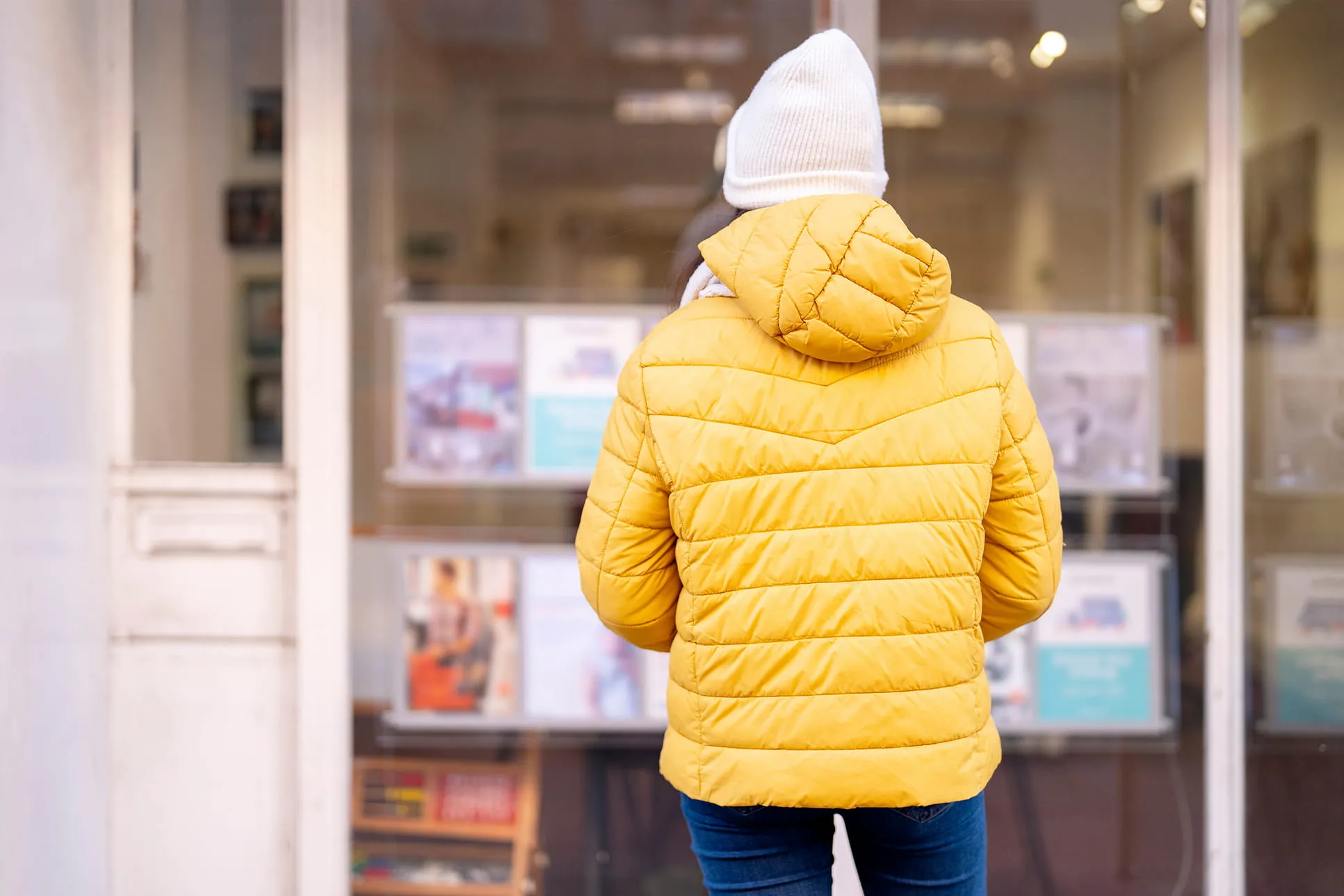 Person in yellow jacket and white knit hat looking at property listings in a real estate agency window.