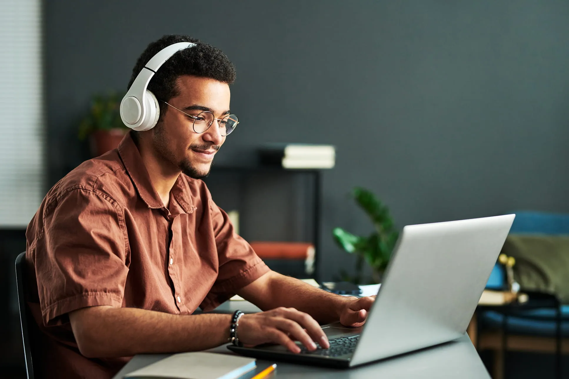 Man with glasses and white headphones working on a laptop at a desk indoors.