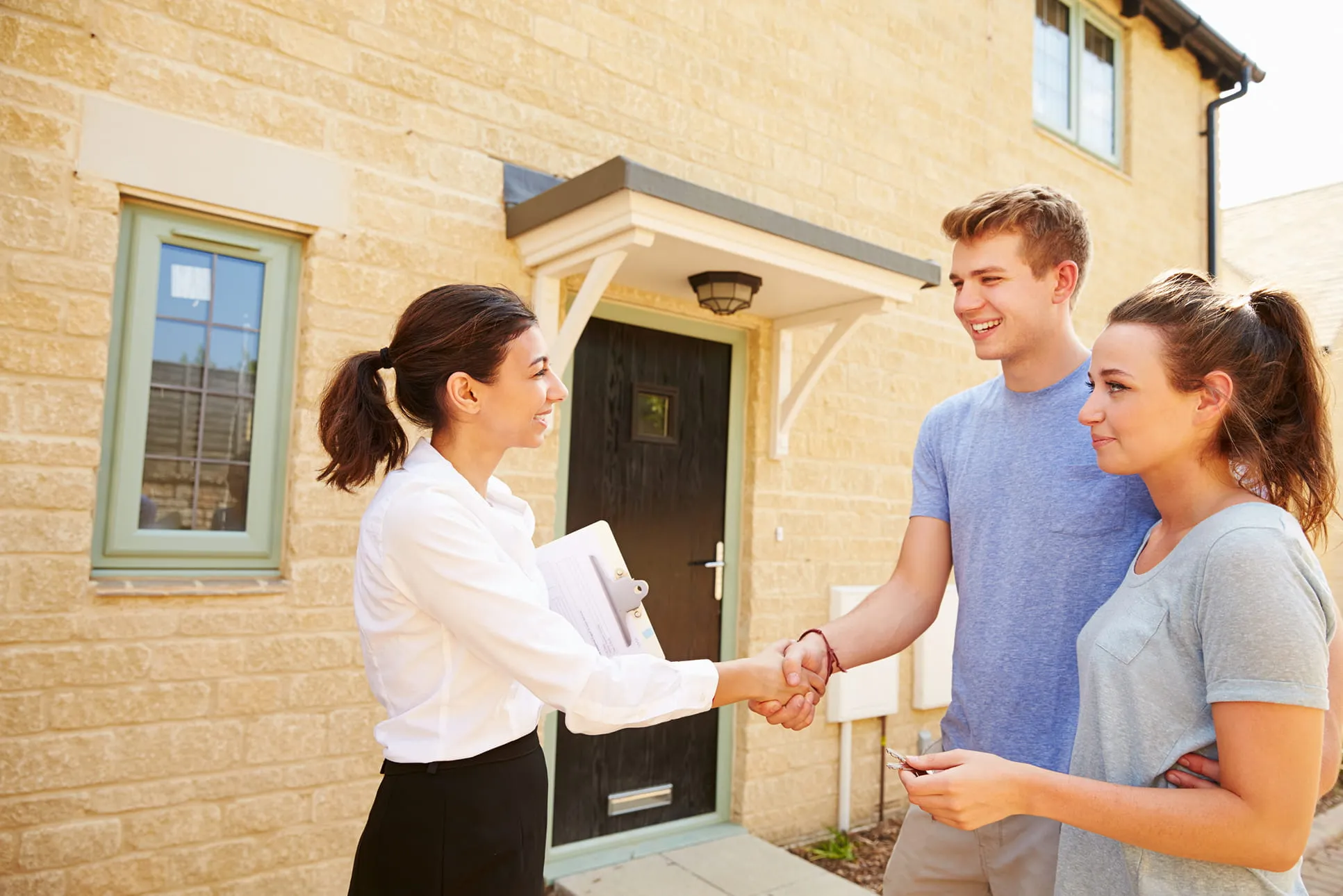 Real estate agent shaking hands with a smiling young couple outside a stone house entrance.