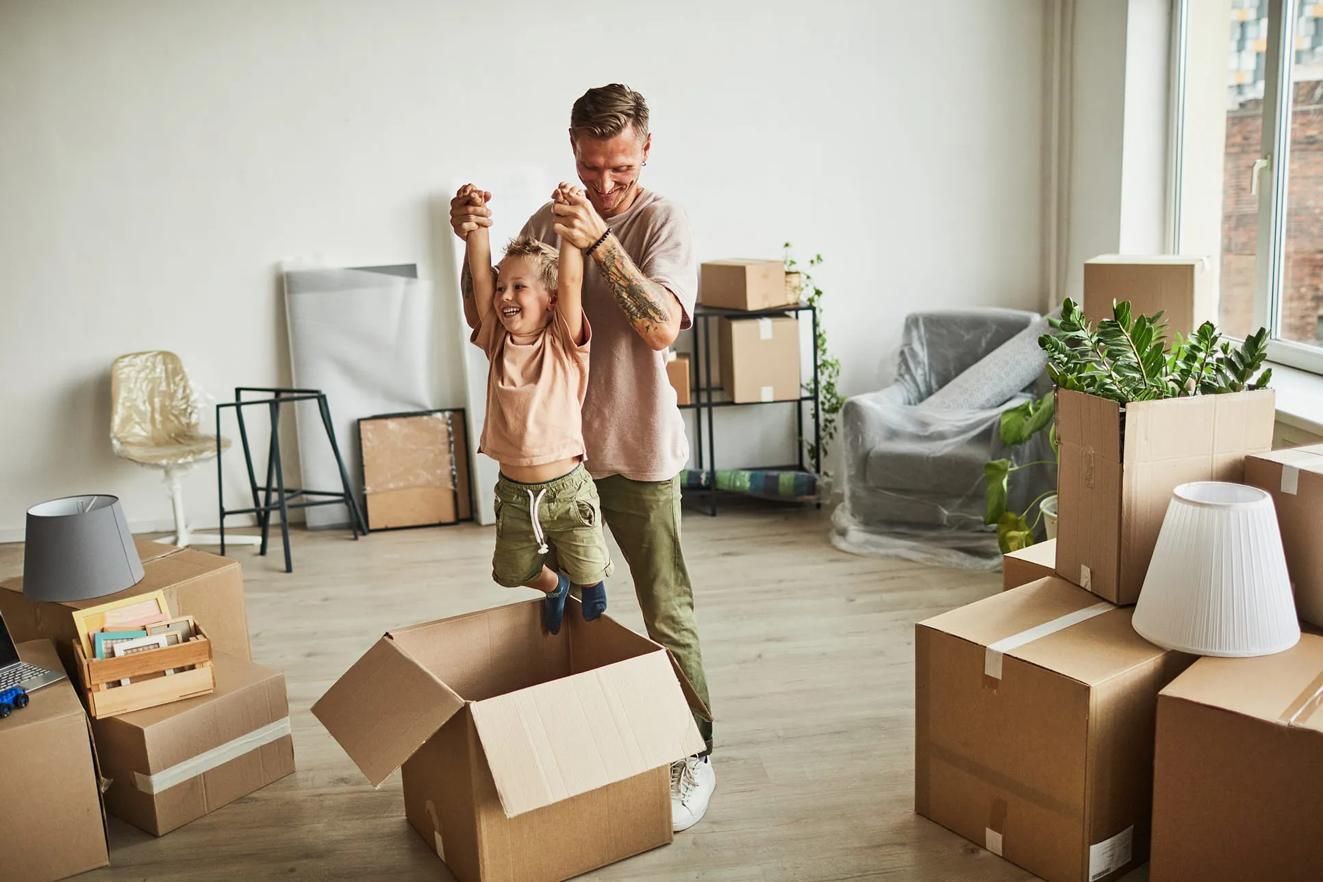 Smiling father lifting his happy young son out of an open cardboard box in a room filled with moving boxes and furniture.