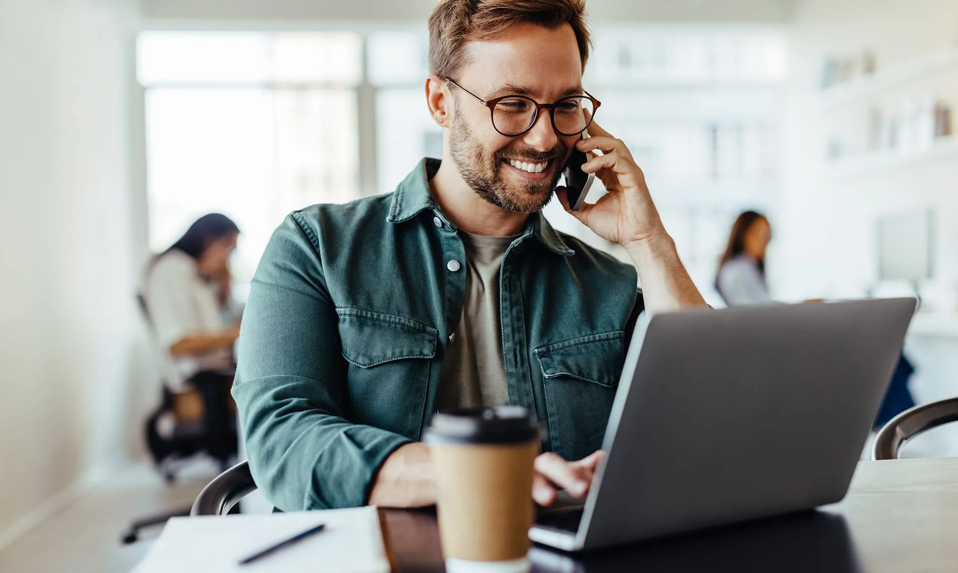 Smiling man wearing glasses talking on a phone while using a laptop at a desk with a coffee cup in an office setting.