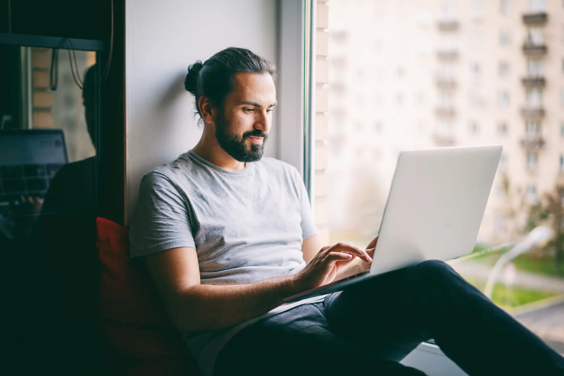 Man with a beard and bun sitting by a window using a laptop.