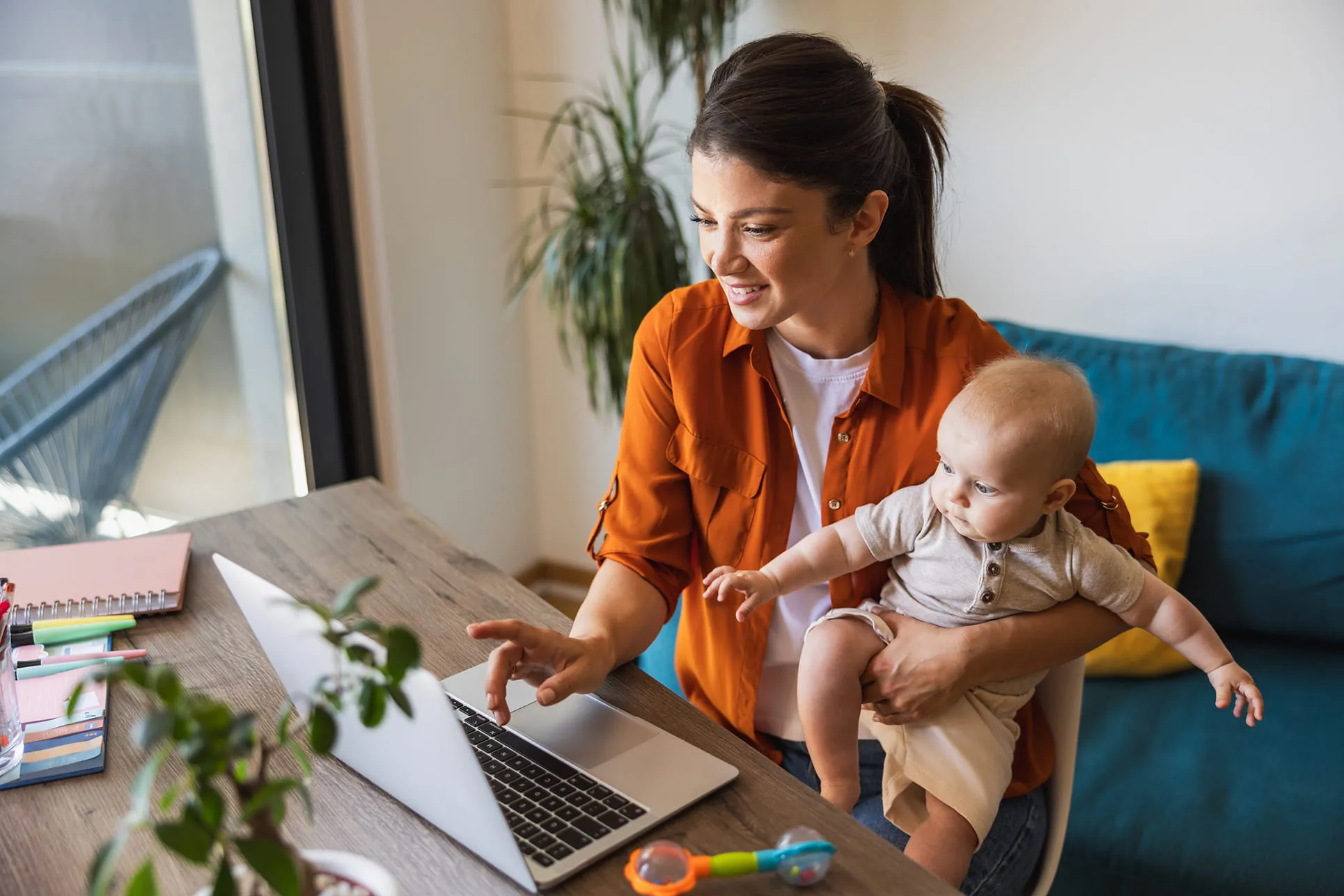 Woman holding baby on her lap while working on a laptop at a wooden desk near a window.