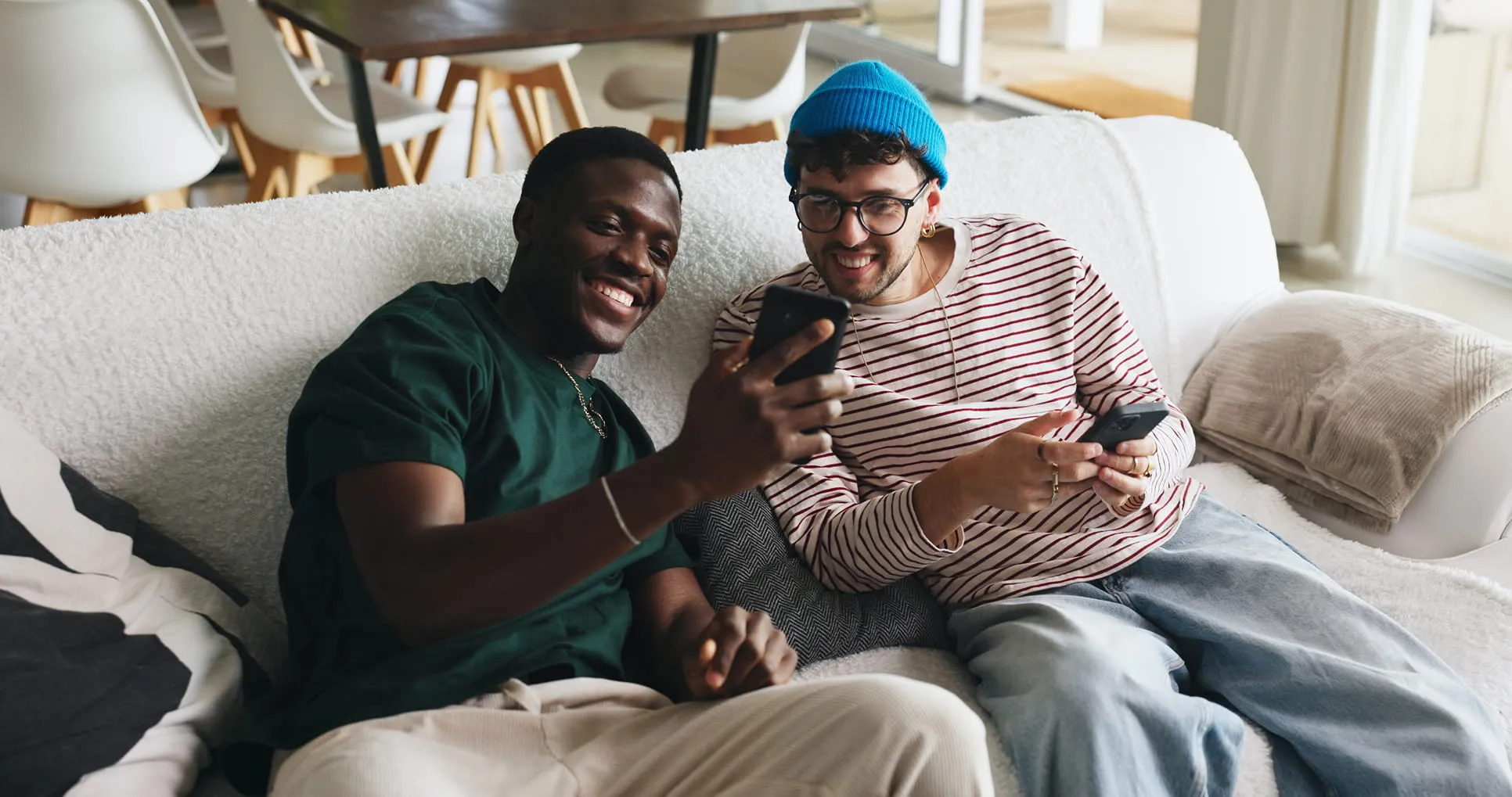Two men sitting on a white couch smiling and looking at their smartphones together.