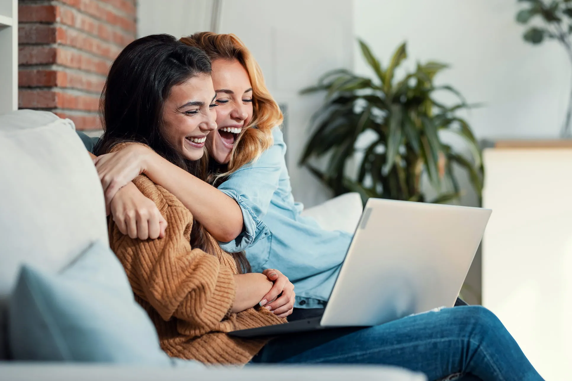 Two smiling women sitting on a couch hugging each other while looking at a laptop.