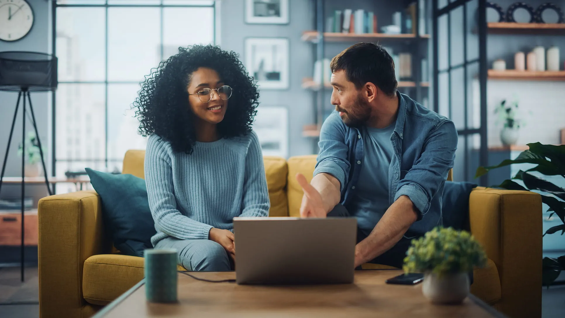 Man and woman sitting on a yellow couch, engaging in a discussion with a laptop on the table in front of them.