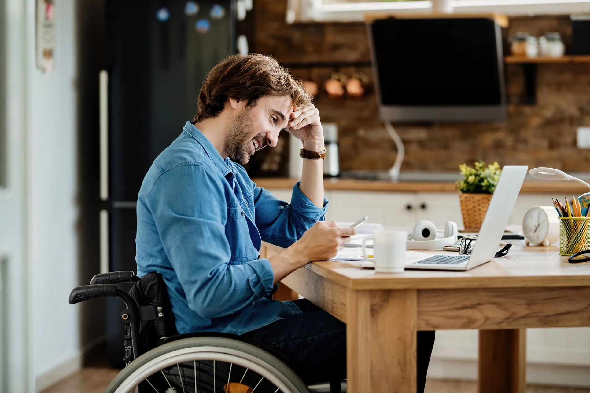Man in wheelchair smiling while looking at his smartphone at a wooden desk with a laptop and coffee mug in a kitchen setting.