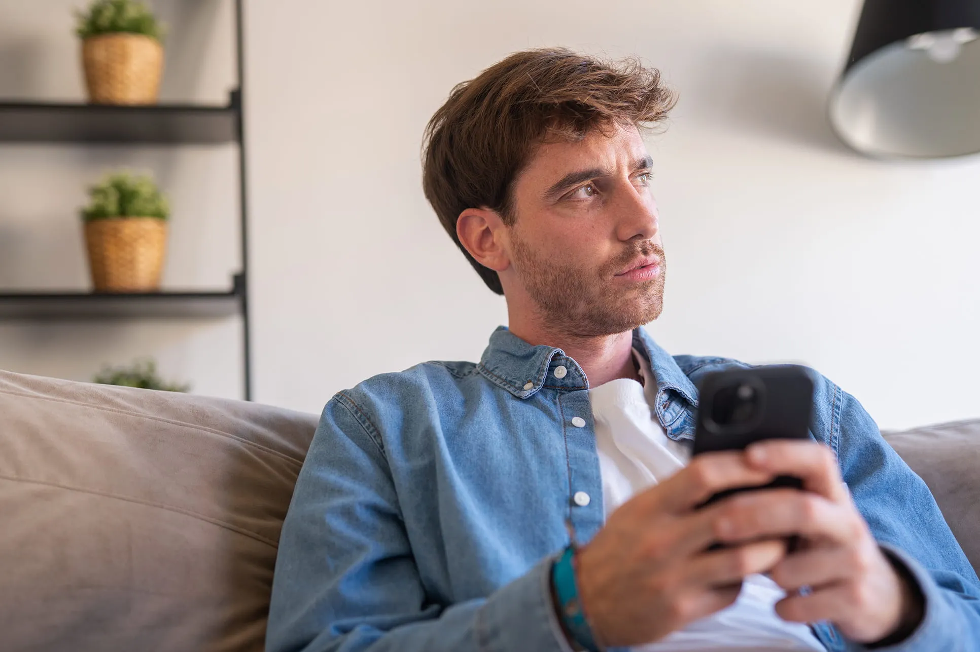 Thoughtful young man in denim shirt holding a smartphone while sitting on a sofa indoors.