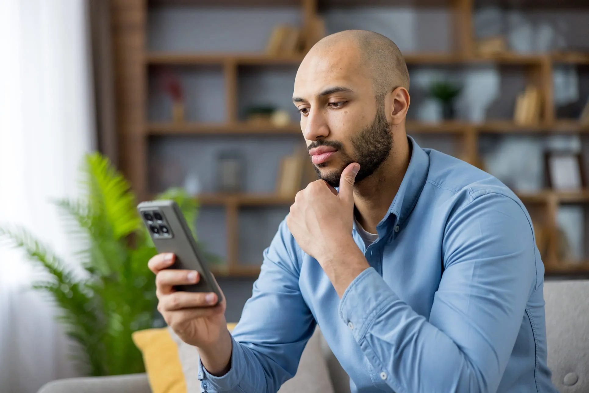 Man in blue shirt sitting on a couch, thoughtfully looking at his smartphone.