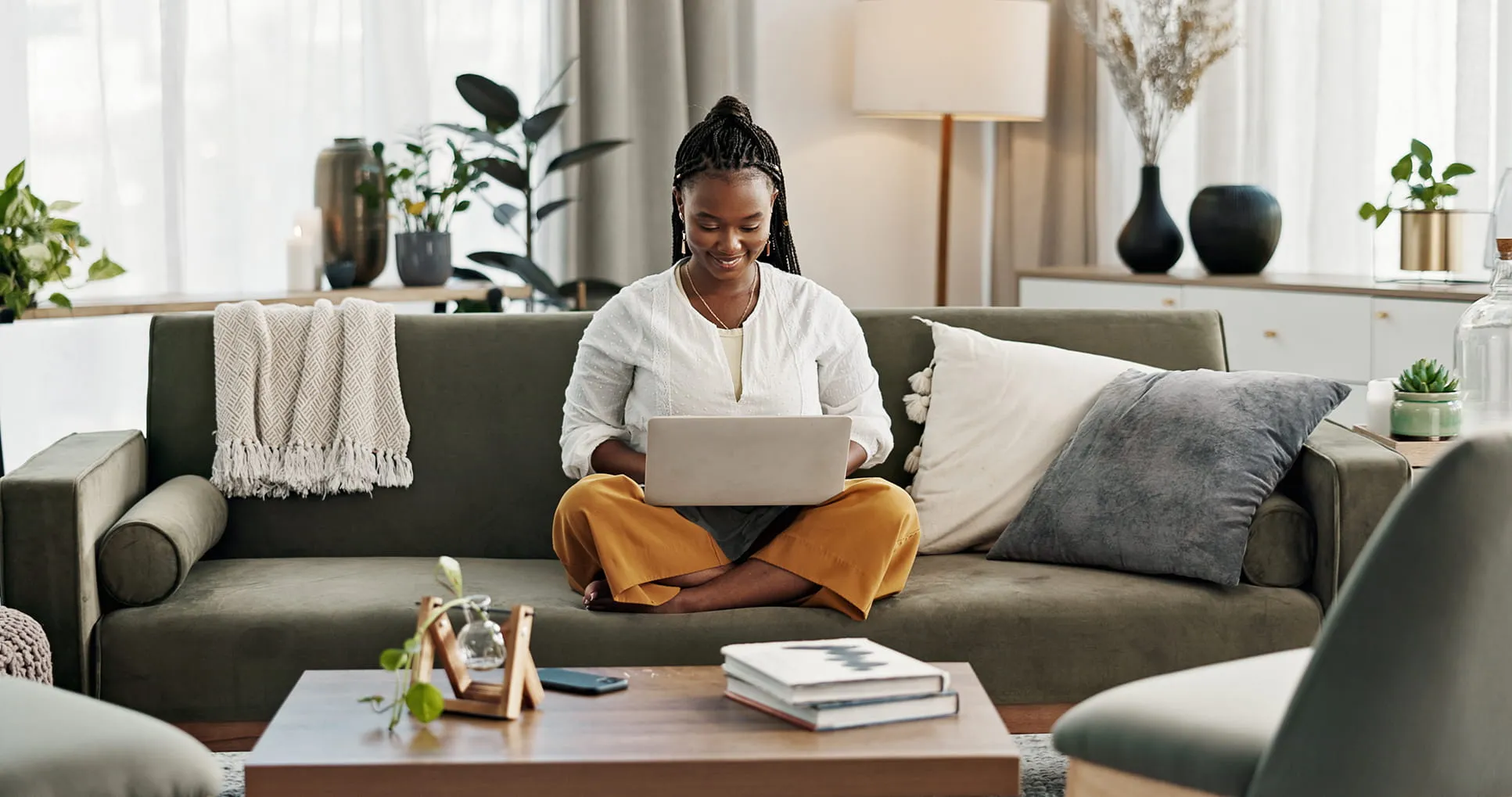 Smiling woman with braided hair sitting cross-legged on a green sofa using a laptop in a cozy living room.