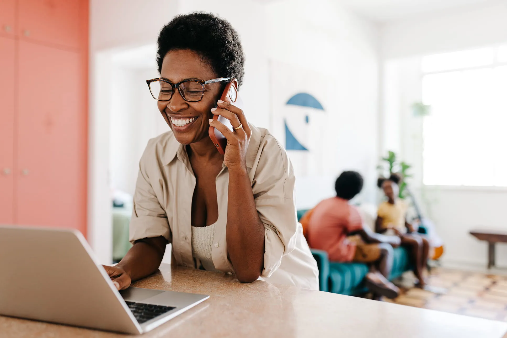 Smiling woman wearing glasses talking on a phone while using a laptop at home, with two children sitting on a couch in the background.