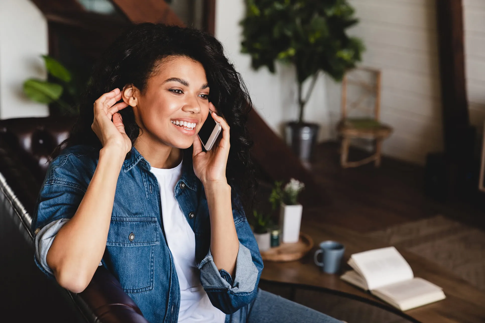 Smiling woman in a denim jacket sitting on a leather sofa while talking on a smartphone.