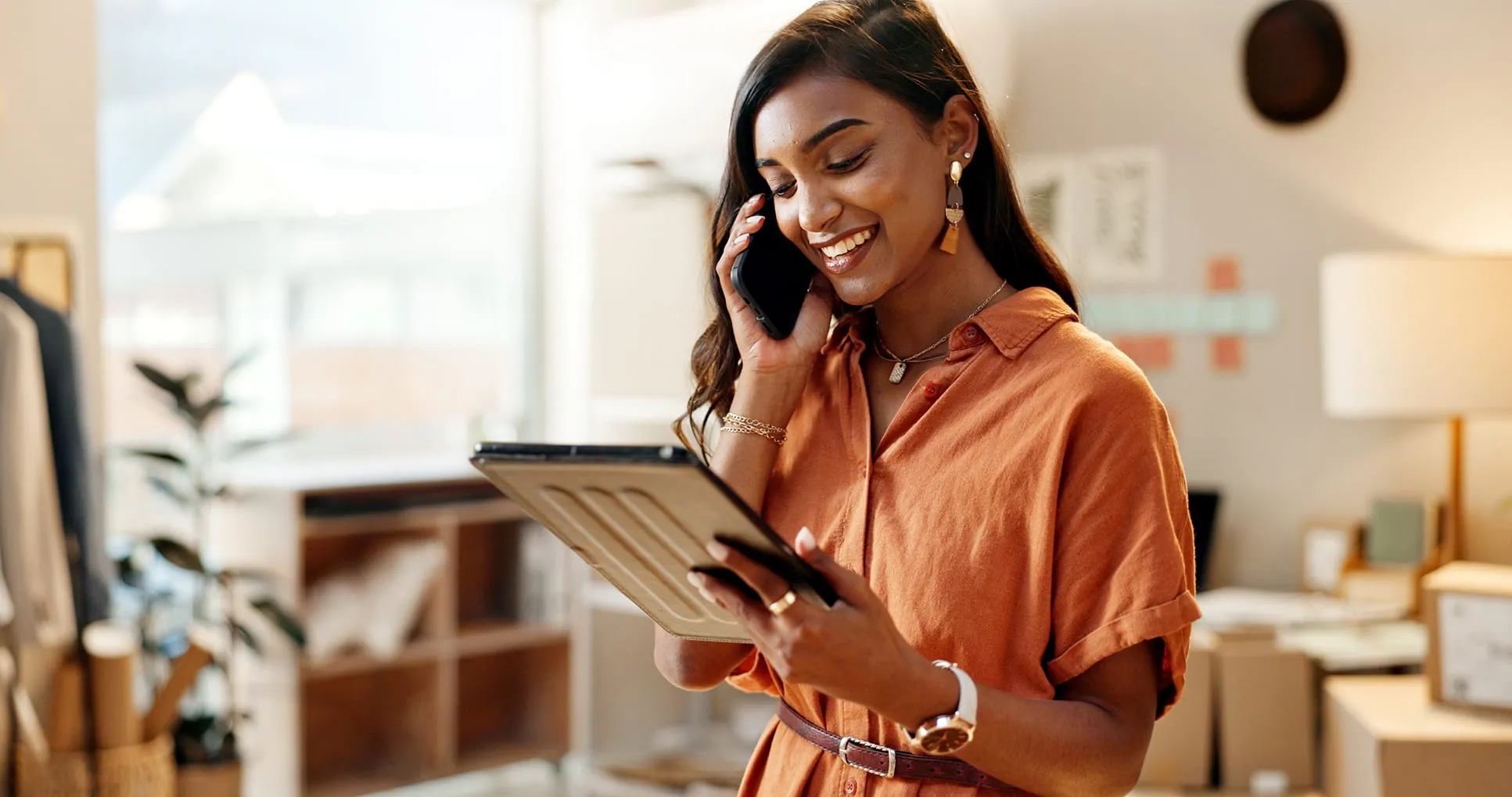 Smiling woman in an orange dress talking on a phone while holding a tablet in a bright room.