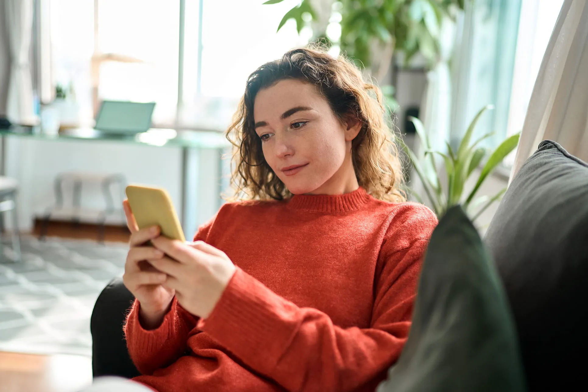 Young woman in a red sweater sitting on a couch using a yellow smartphone in a bright living room.