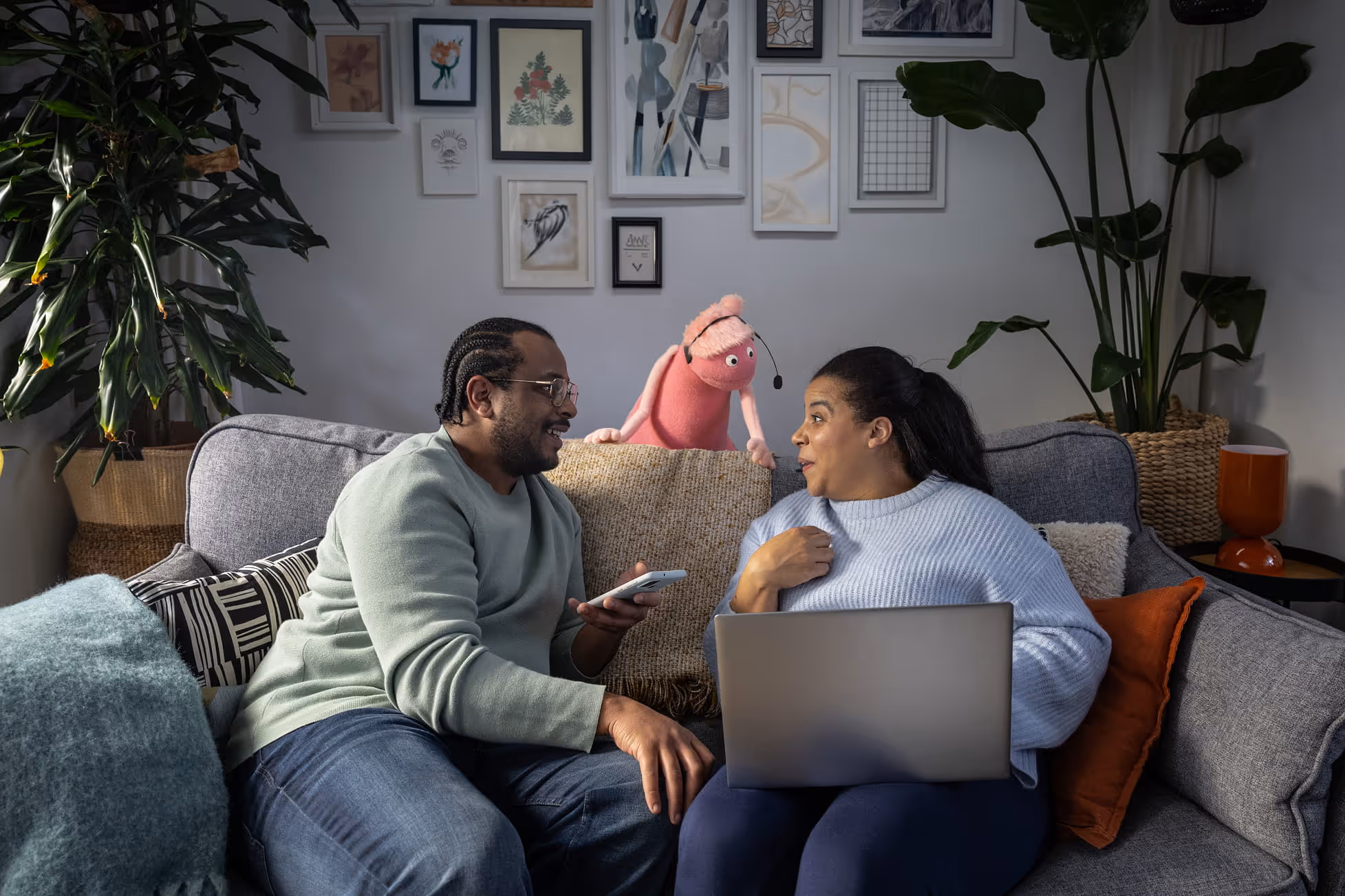 Man and woman sitting on a couch talking with a pink puppet wearing a headset behind them.