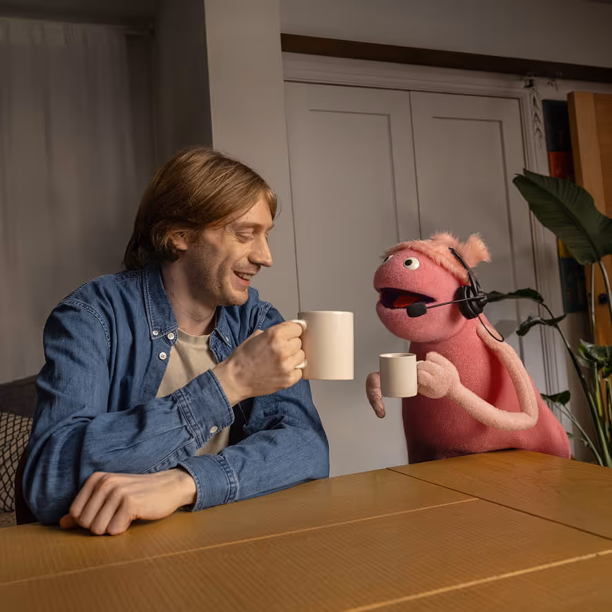 Man in denim shirt smiling and clinking coffee mugs with a pink puppet wearing a headset at a table.