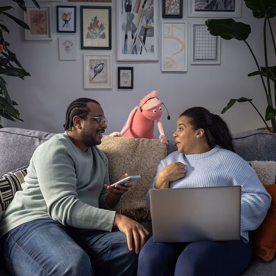 Man and woman sitting on a couch with a laptop and phone, interacting with a pink puppet wearing a headset.