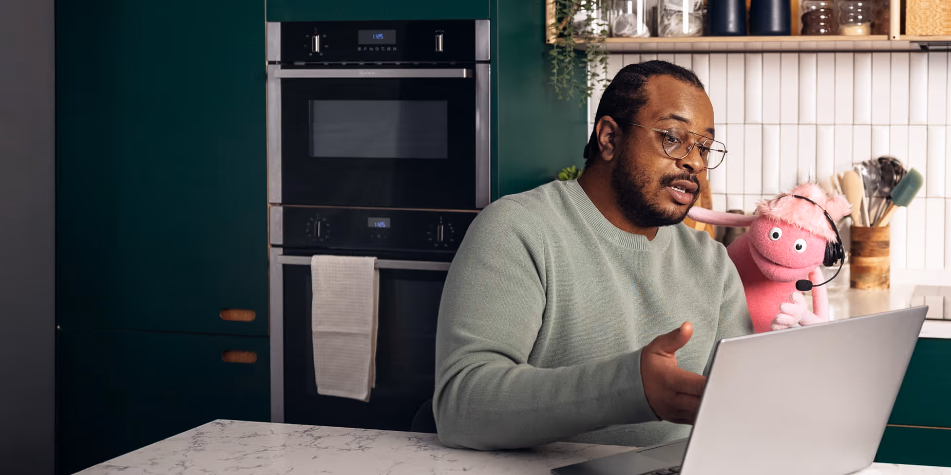 Man wearing glasses sitting at a kitchen counter working on a laptop next to a pink puppet with a headset.