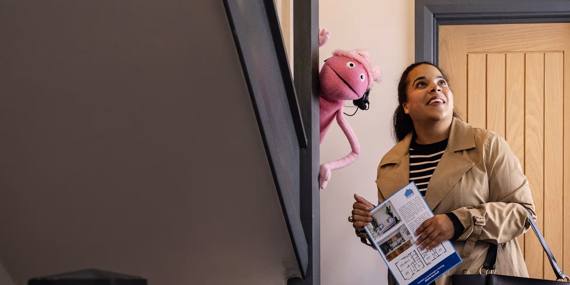 Woman holding a floor plan brochure looking up while a pink puppet with headphones peeks around a door.