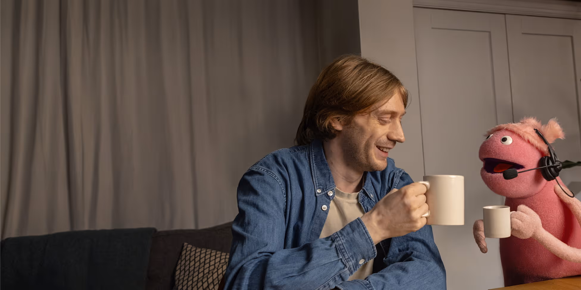 Man in denim shirt smiling and clinking coffee mugs with a pink puppet wearing a headset.
