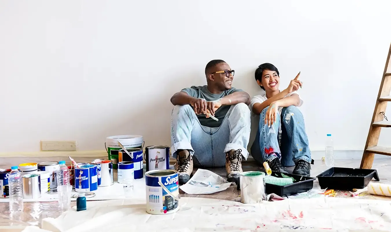 A smiling couple sitting on the floor against a white wall, surrounded by paint cans, brushes, and painting supplies, taking a break from painting.