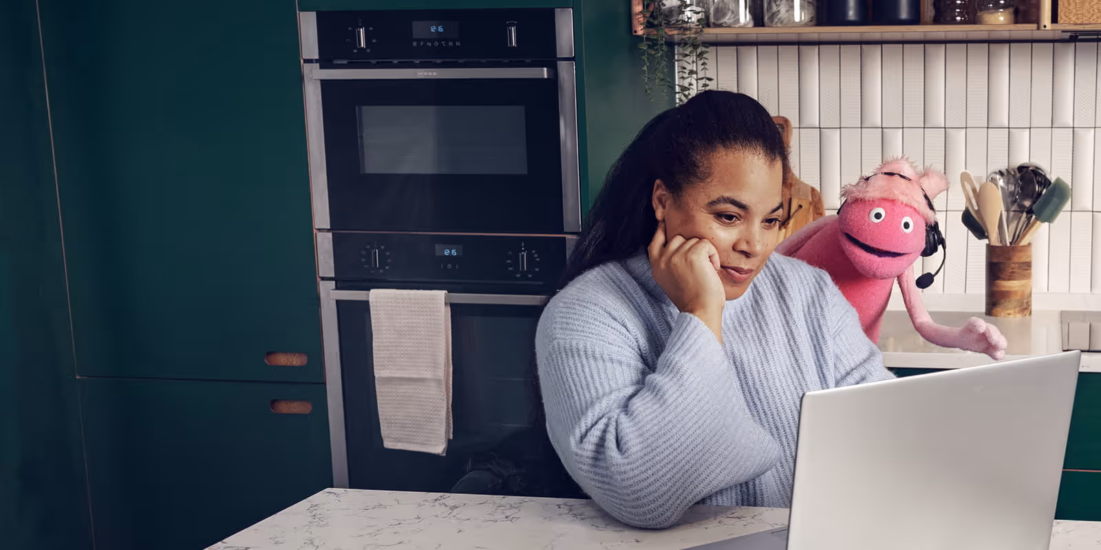 Woman in a blue sweater looks at a laptop in a kitchen with green cabinets, while a pink puppet with a headset stands behind her.