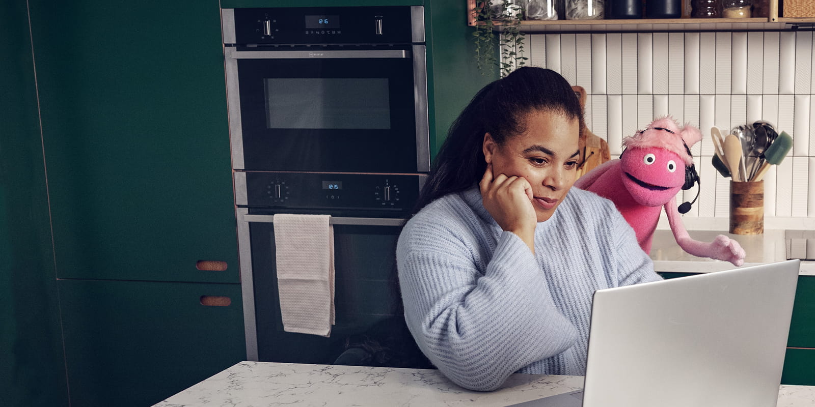 Woman in a blue sweater looks at a laptop in a kitchen with green cabinets, while a pink puppet with a headset stands behind her.
