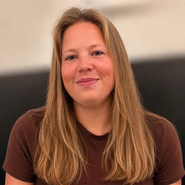 Smiling woman with long blonde hair wearing a brown shirt against a blurred beige and black background.